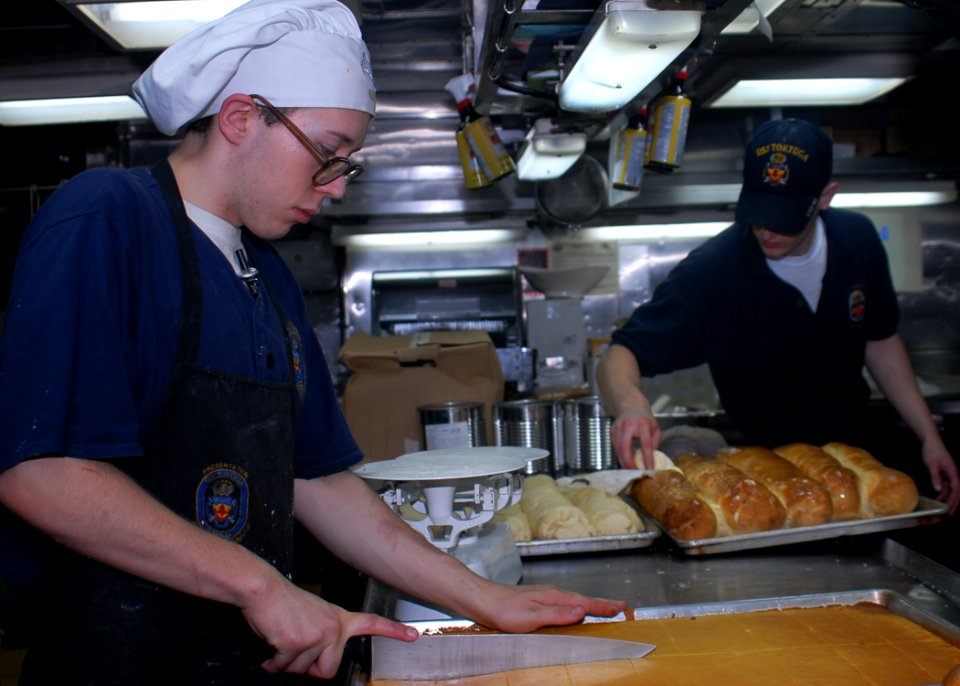 US Navy 091104-N-6692A-085 Culinary Specialist Seaman Levi Chute, left, cuts cornbread into portions while Culinary Specialist Seaman Shawn Carlile prepares loaves of bread for dinner aboard the amphibious dock landing ship USS