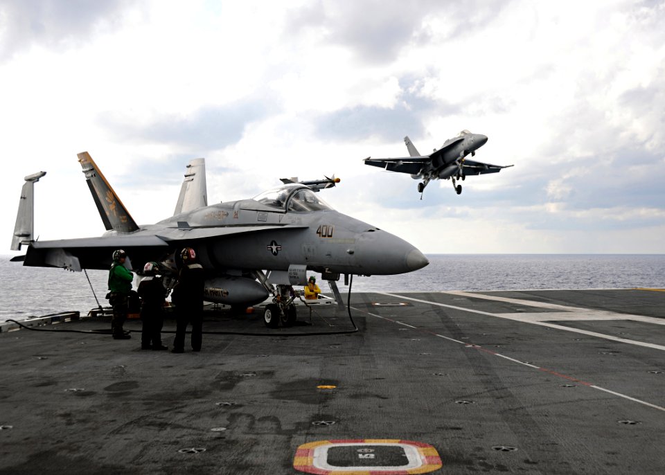 US Navy 100521-N-3885H-193 An F-A-18C Hornet assigned to Strike Fighter Squadron (VFA) 15 approaches the aircraft carrier USS George H.W. Bush (CVN 77) for a landing while an F-A-18 Hornet assigned to Strike Fighter Squadron (V
