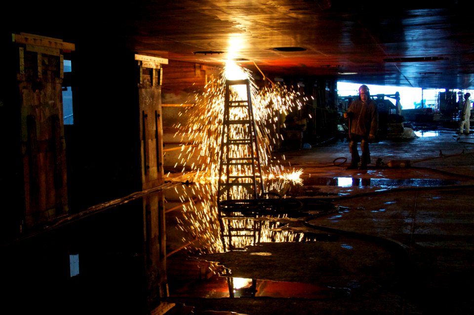 US Navy 101221-N-9643K-440 A NASSCO shipyard worker stands fire watch as work continues beneath the hull of the amphibious assault ship USS Bonhomm