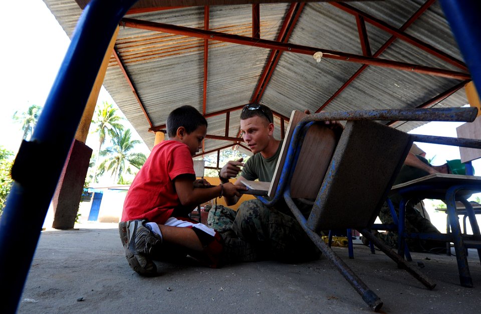 US Navy 110128-N-7589W-405 Cpl. Paul Fuit, assigned to 2nd Marine Logistics Group, shows a child how to use a rasp to smooth wood desks at Escuela