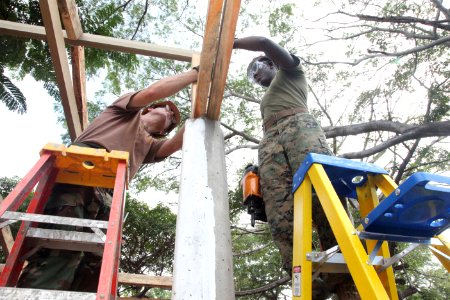 US Navy 111212-A-IP644-113 Builder 2nd Class Phillip Grindstaff and LCpl. Ronald Russell use a nail gun to steady rafters on an outdoor classroom b