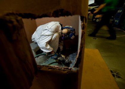 US Navy 120124-N-DR144-419 Airman Recruit Travis Nesbit works inside a cardboard shipping container to control dust and flying debris as he sands o