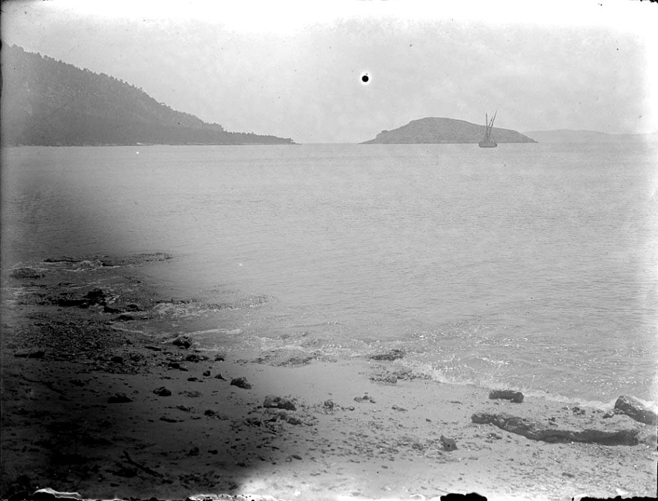 Vista d'una platja amb un vaixell i muntanyes al fons (AFCEC RIBASV C 2679)
