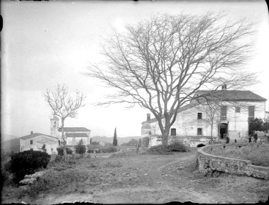 Vista del camí d'un poble amb un arbre al mig, gent i un campanar al fons (AFCEC RIBASV C 2521)