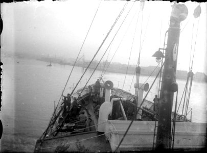 Vista parcial de la coberta d'un barc navegant amb homes treballant i un port al fons (AFCEC RIBASV C 2660)