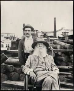 Walt Whitman, three-quarter length portrait, seated, facing front, with his nurse Warren Fritzenger, standing, and a pile of logs behind him, on the wharf, probably near his Mickle Street LCCN97506894