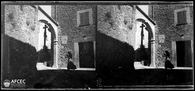 Una dona caminant per un carrer de Pollença amb un arc i una creu de terme (AFCEC VINTRO B 10543)