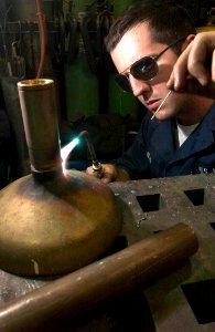 US Navy 060902-N-9851B-008 Machinery Repairman 2nd Class Timothy Millay brazes a brass pipe fitting to the bell of a brass decontamination shower head, in the general workshop aboard the guided missile destroyer USS Hopper (DDG photo
