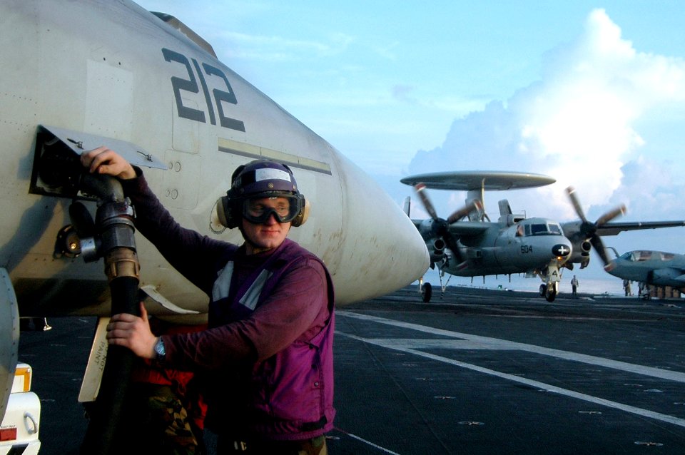 US Navy 060909-N-8604L-105 Airman Douglas Riley fuels an F-A-18 Hornet assigned to Strike Fighter Squadron Two Seven (VFA-27) as an E-2C Hawkeye from Carrier Airborne Early Warning Squadron One One Five (VAW-115) lands