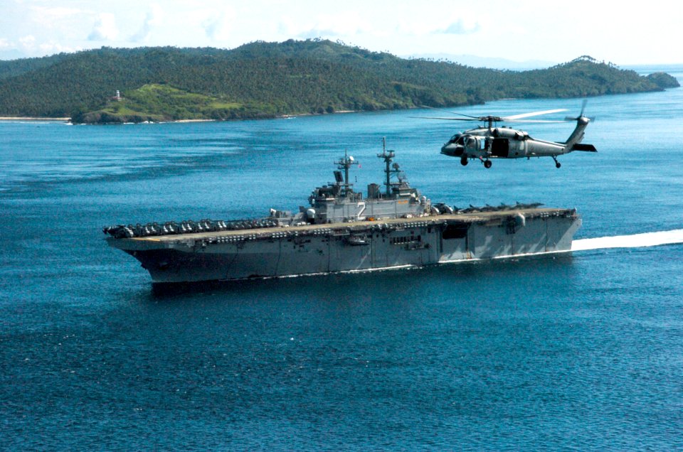 US Navy 061104-N-5067K-064 With a MH-60S Seahawk assigned to the Island Knights of Helicopter Sea Combat Squadron Two Five (HSC-25) flying as a channel guard, USS Essex (LHD 2) passes the Capul Island Lighthouse