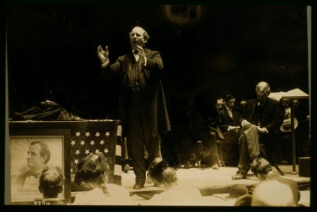 William Jennings Bryan, full-length view standing on stage, delivering campaign speech, another man seated to the rear of the stage LCCN2001697075