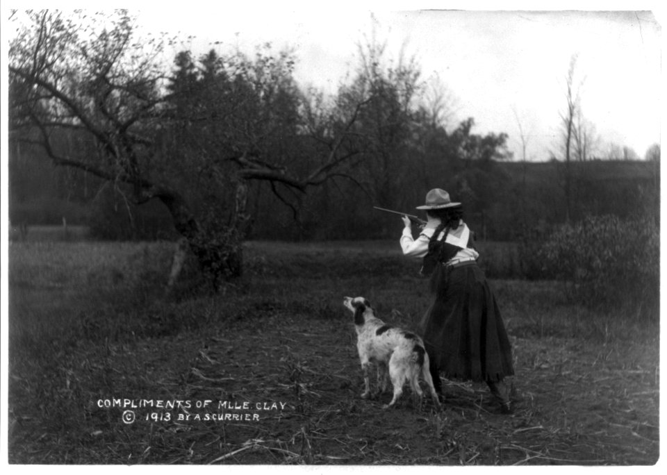 Woman in cowgirl outfit aiming small rifle; English Setter standing beside her LCCN2005680972