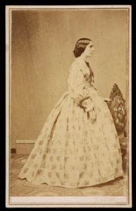 Woman in floral dress, standing by chair, facing right LCCN2016653154