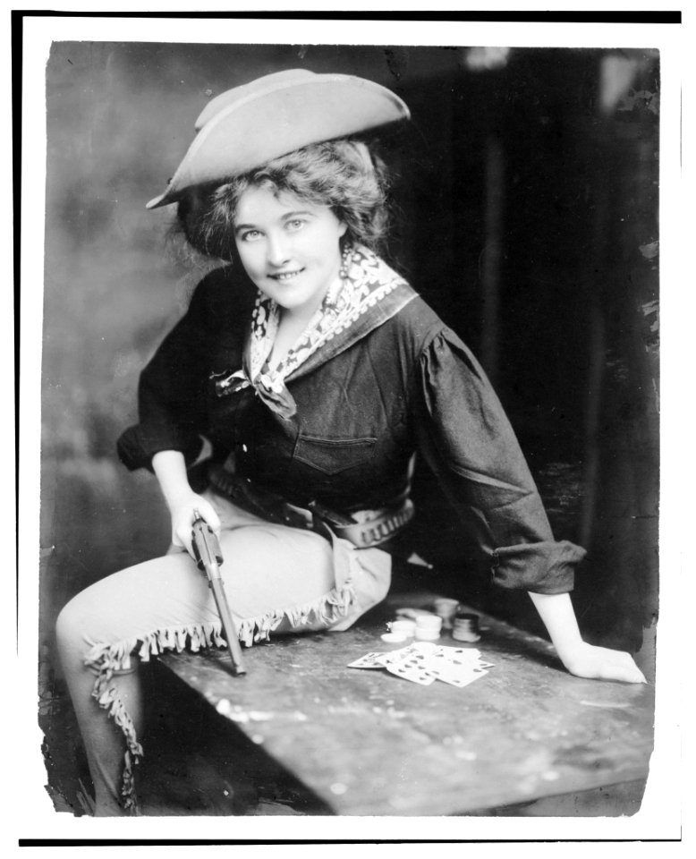 Woman posing in cowgirl outfit, holding revolver, seated on table, with playing cards LCCN93512431