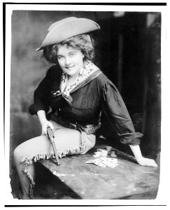 Woman posing in cowgirl outfit, holding revolver, seated on table, with playing cards LCCN93512431