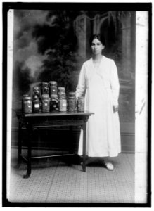 Woman standing next to table holding a display of preserved food in glass jars, including peas, beans, fruit, and eggs LOC hec.11490