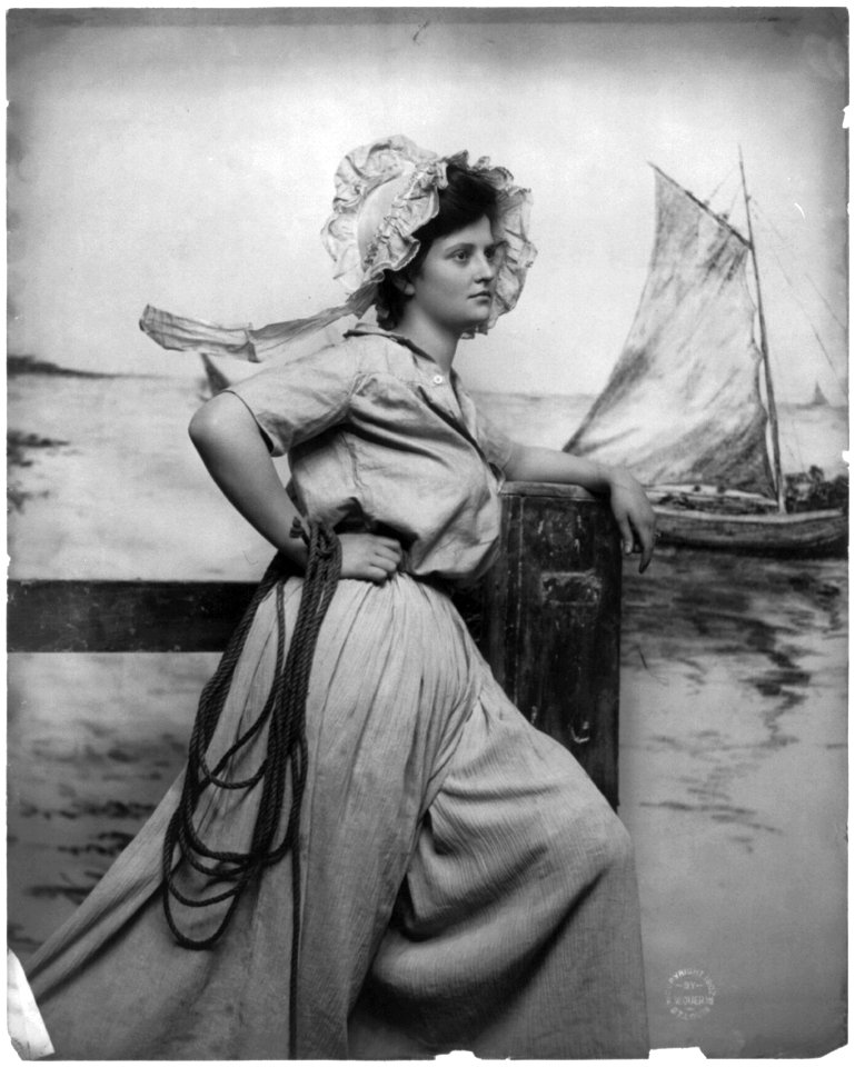 Woman wearing bonnet, holding rope and standing on pier; fishing boat in background LCCN2003677204