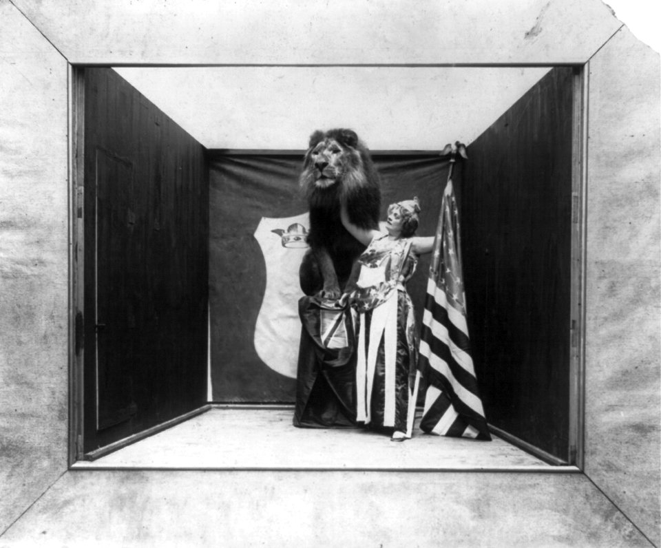 Woman with stars and eagle on her costume holding U.S. flag and touching tame lion under its neck LCCN2002711737