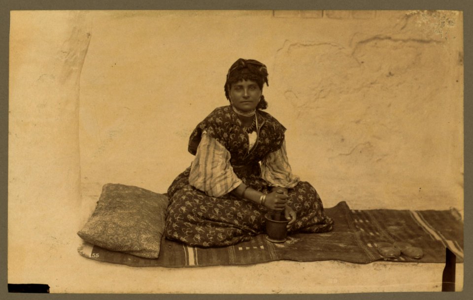 Woman, full-length portrait, seated on rug on ground, facing front, using mortar and pestle, Constantine, Algeria) - ND LCCN2004666995