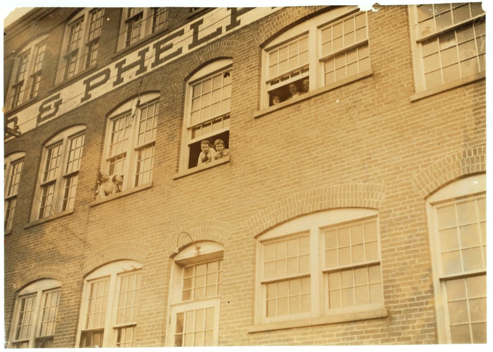 Workers in Fargo & Phelps Shoe Co., Saw six boys and two girls probably under 14. Note face of very young girl in window. Large sign up, 'Boys Wanted.' LOC nclc.04711
