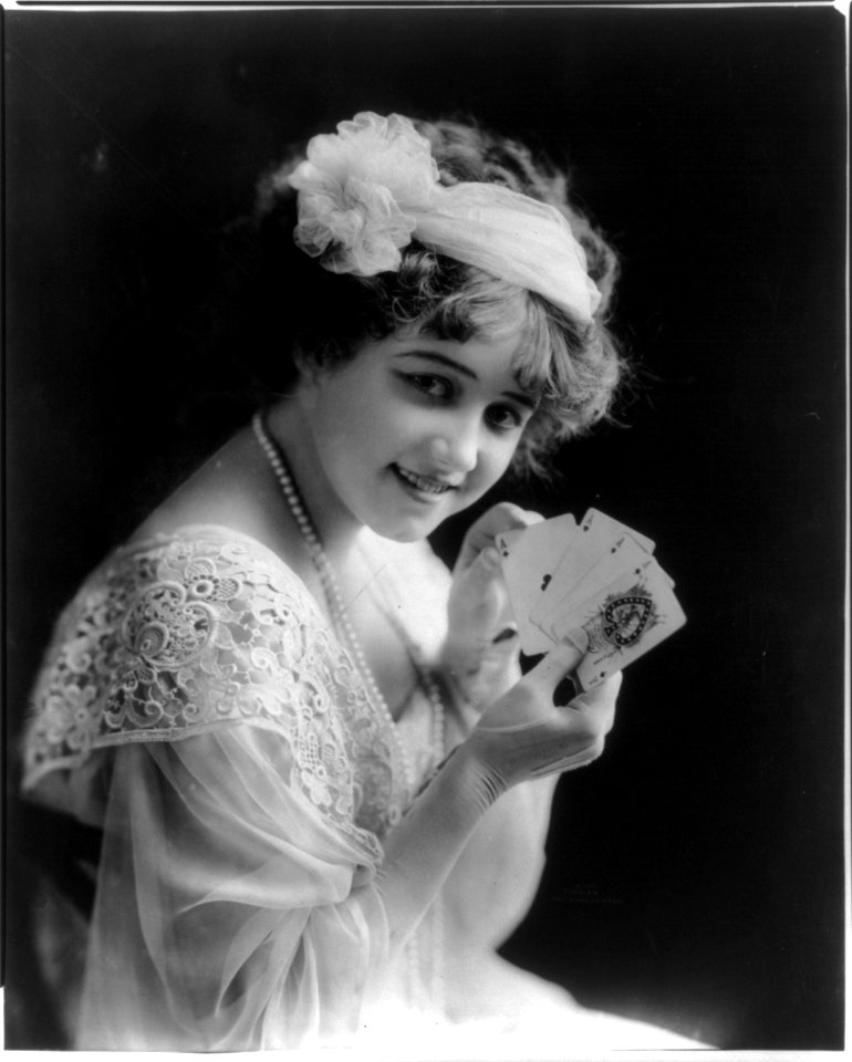 Young woman, half-length portrait, seated, facing front, wearing lace dress and head wrap holding four aces LCCN2012646441