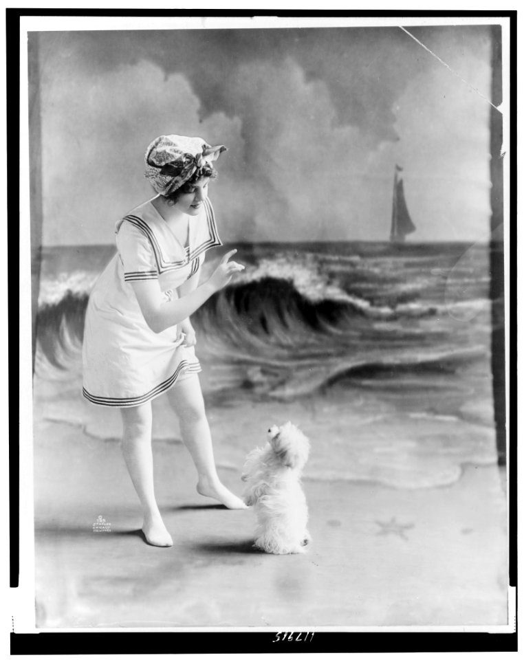 Young woman in bathing suit with small dog in front of studio backdrop of beach LCCN97513783