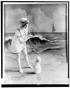 Young woman in bathing suit with small dog in front of studio backdrop of beach LCCN97513783