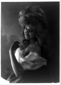 Young woman modeling- half lgth., facinag left, wearing low-cut dress and hat with flowers LCCN2003677217