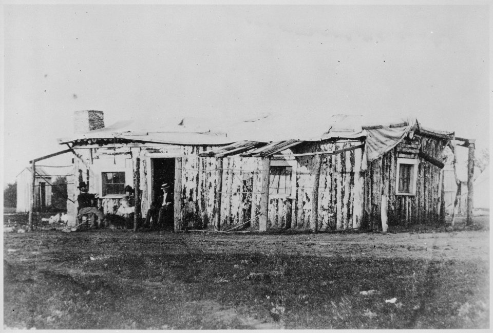 Officers quarters at Ft. Rawlins, Wyoming, May 7, 1877. Two men and a woman holding a baby sit in front of the crude s - NARA - 531107