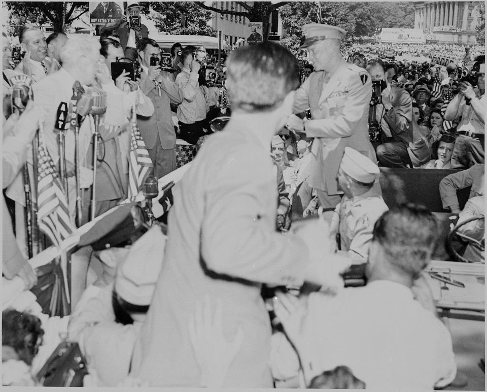 Photograph of General Dwight D. Eisenhower standing in the back of his jeep, surrounded by photographers and cheering... - NARA - 199113