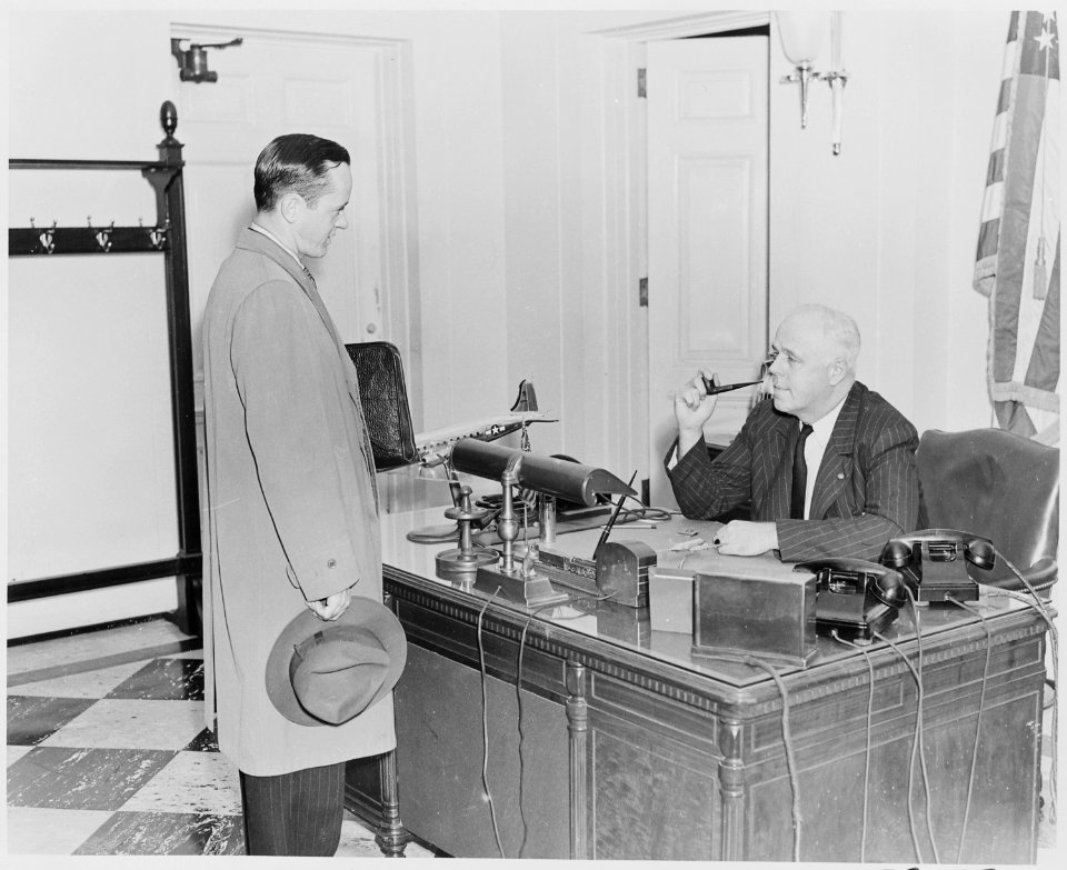 Photograph of William Simmons, receptionist at the White House, seated at his desk talking with an unidentified visitor. - NARA - 199456