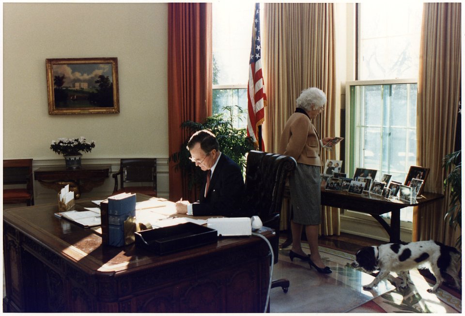 President Bush works at his desk in the Oval Office as Mrs. Bush looks at photographs on the table behind the Oval... - NARA - 186389