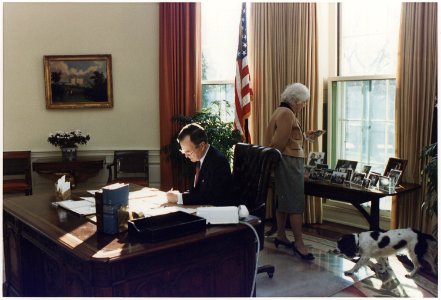 President Bush works at his desk in the Oval Office as Mrs. Bush looks at photographs on the table behind the Oval... - NARA - 186389 photo