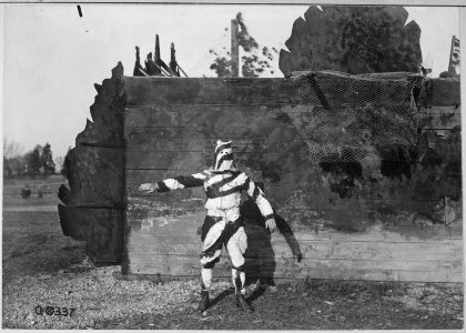 Soldier in black and white uniform to conceal him while climbing trees. He stands in front of a house camouflaged to... - NARA - 530710