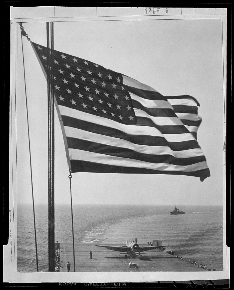 The Stars and Stripes wave over a U.S. Navy aircraft carrier at sea. On this flight deck is a Grumman Avenger TBM... - NARA - 520695