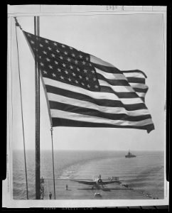 The Stars and Stripes wave over a U.S. Navy aircraft carrier at sea. On this flight deck is a Grumman Avenger TBM... - NARA - 520695