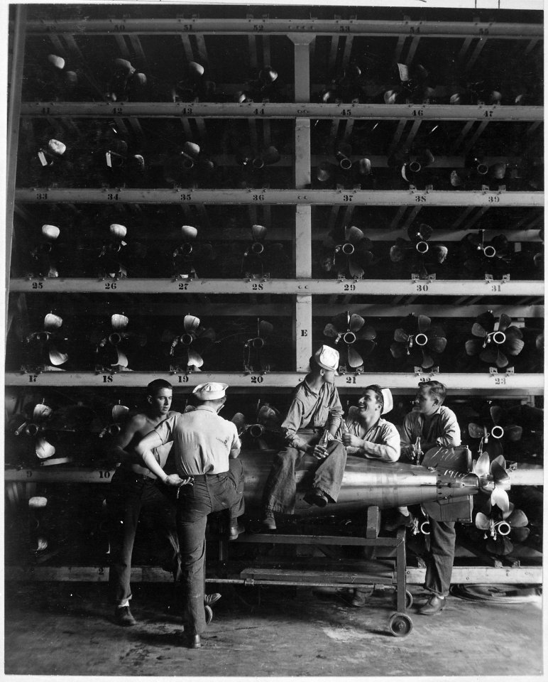Torpedomen relaxing beneath rows of deadly torpedoes in torpedo shop. - NARA - 520832