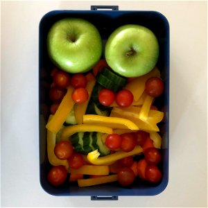 A lunch box filled with two granny smith apples and a selection of fresh vegetables, like cherry tomatoes, bell pepper, cucumber and carrots. photo